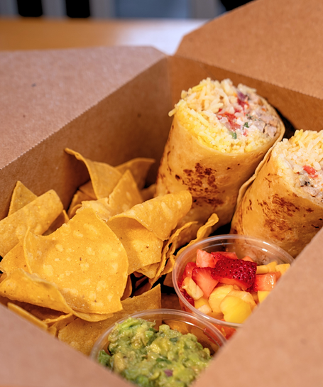 A cardboard box with a burrito, tortilla chips, fruit, and guacamole